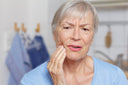 An elderly lady with a blue top standing in the kitchen holding her mouth looking in slight pain
