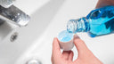 A man's hand holding a mouth rinse cap over a white sink and filling the cap with blue mouth rinse from the other hand