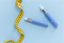 Two blue weight loss injections next to a yellow tape measure on a light blue background