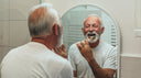 An older male with white hair brushing teeth looking in a arched mirror