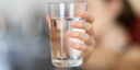A glass of water in focus being held by a lady's hand blurred in the background